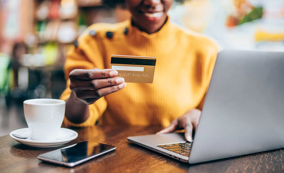 A woman in yellow holding a credit card while working on a laptop in a coffee shop.