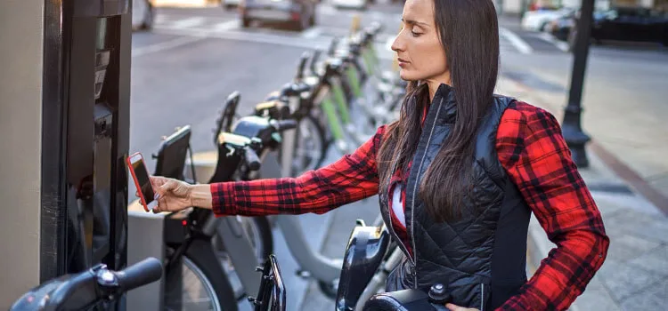 A woman standing outside, holding up her mobile device to an unattended payment machine at a bike stand.