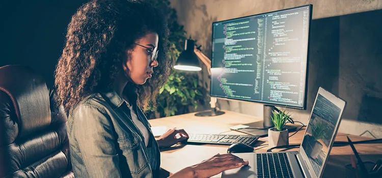 A woman working on a desktop computer and a laptop in dim lighting with coding data on the desktop computer screen.