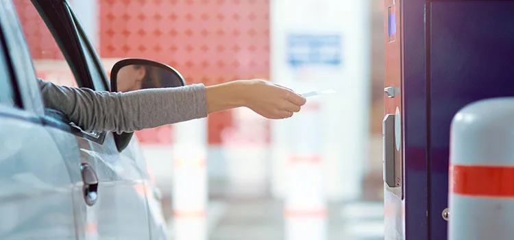 An arm reaching out of a car window to insert a credit card into an unattended payment machine at a parking lot.