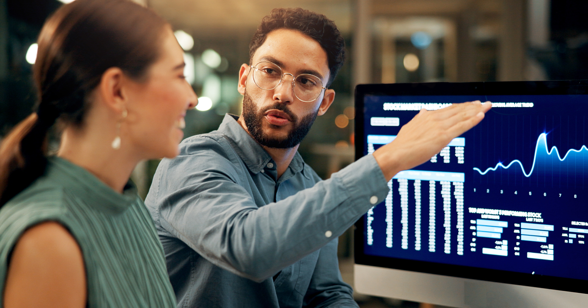 NMI&AI-Blog-Thumbnail Image of man talking to woman in front of a computer. They are talking about AI and how NMI is making the payments experience better for their partners with AI.