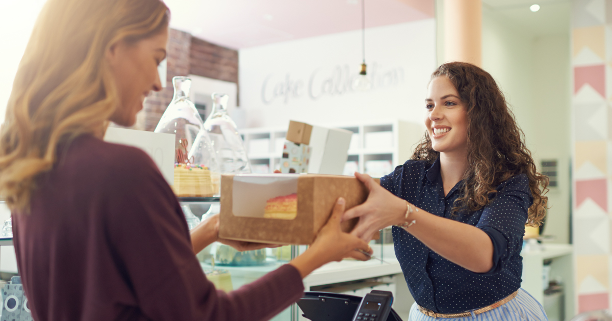 A bakery employee hands a boxed cake to a smiling customer, representing positive in-store shopping experiences. NMI article highlights retail, gifting and payment themes.