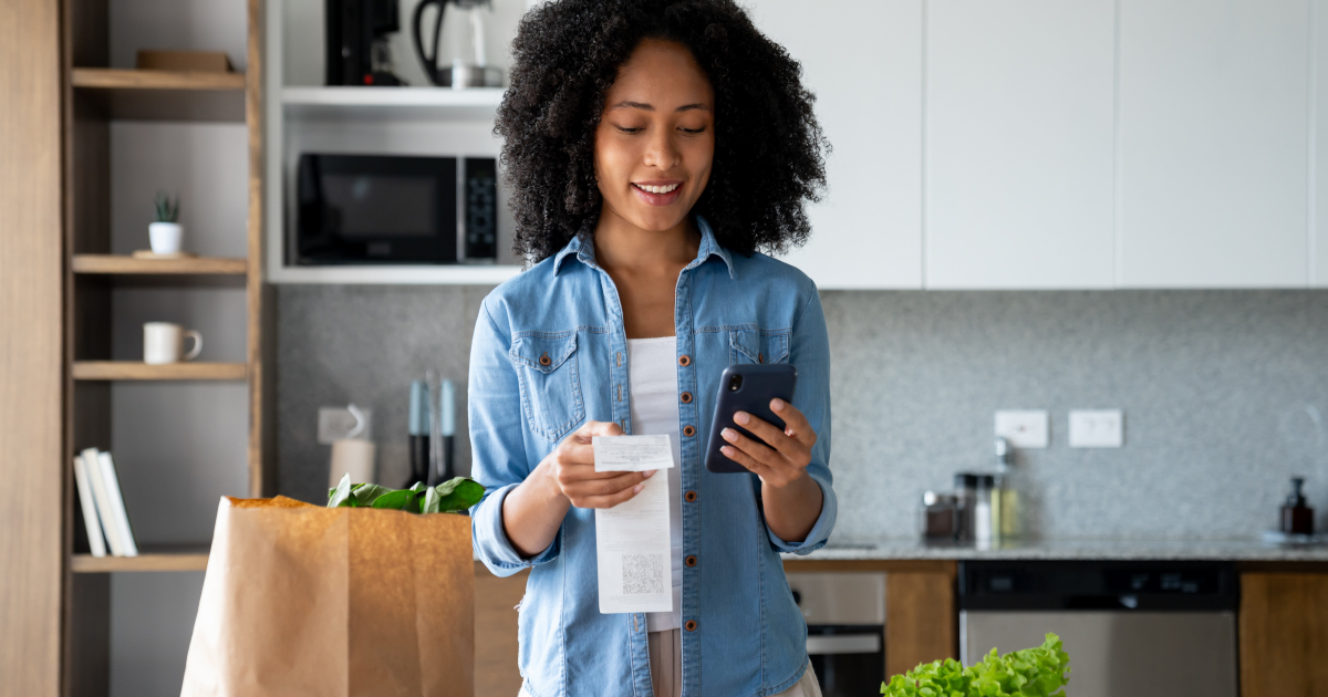 Smiling woman using her smartphone to review a digital receipt in her kitchen, symbolizing smart spending, savings, and modern payment flexibility—representing modular payments in a cost-conscious market.