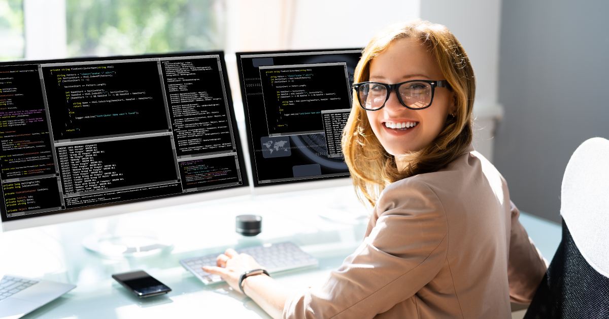 Smiling female developer testing embedded payments in a sandbox environment on dual monitors with coding interfaces, ideal for SaaS platforms.