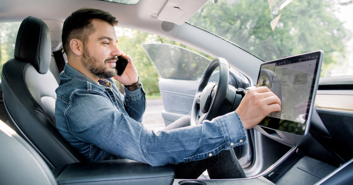 Man using in-car touchscreen interface while talking on the phone, illustrating NMI's prediction of the rise of connected vehicle technology and invisible payments in 2026.