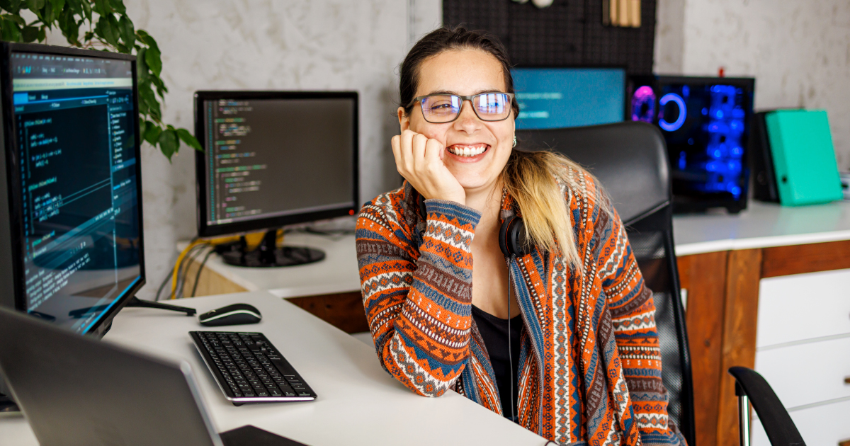 Smiling software developer wearing headphones and glasses, seated at a desk with multiple computer monitors displaying code in a modern workspace.