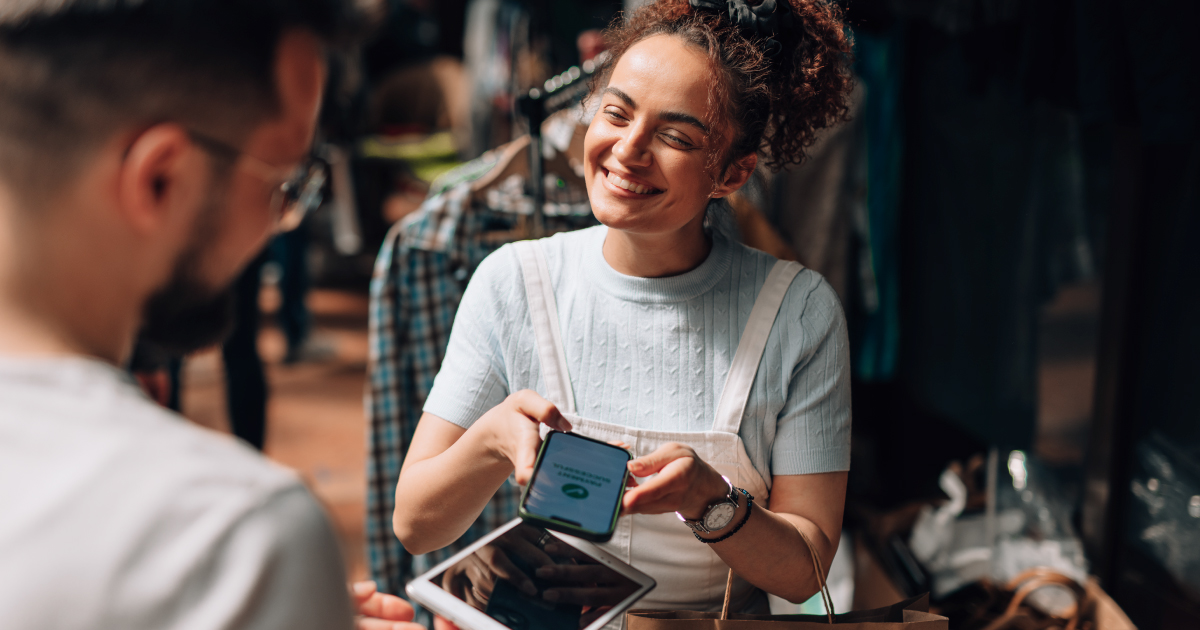 Lady sitting in restaurant smiles as she points her phone to a POS device to tap and pay.