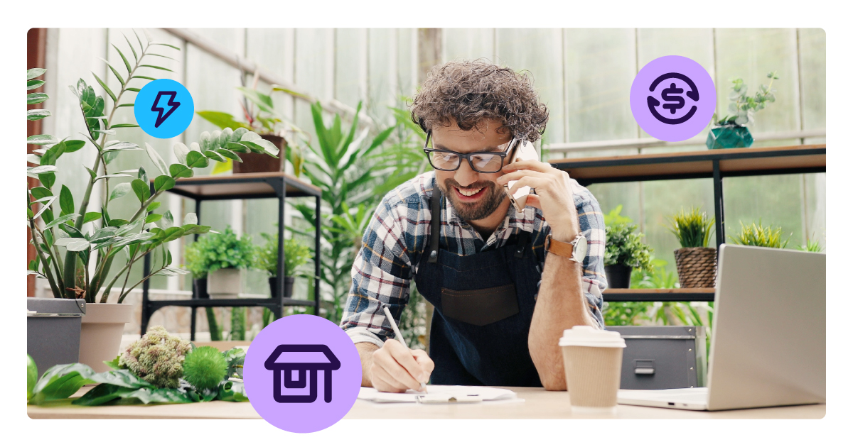 Person wearing glasses and an apron writes on a clipboard while talking on a phone at a desk with a laptop and coffee, surrounded by plants.