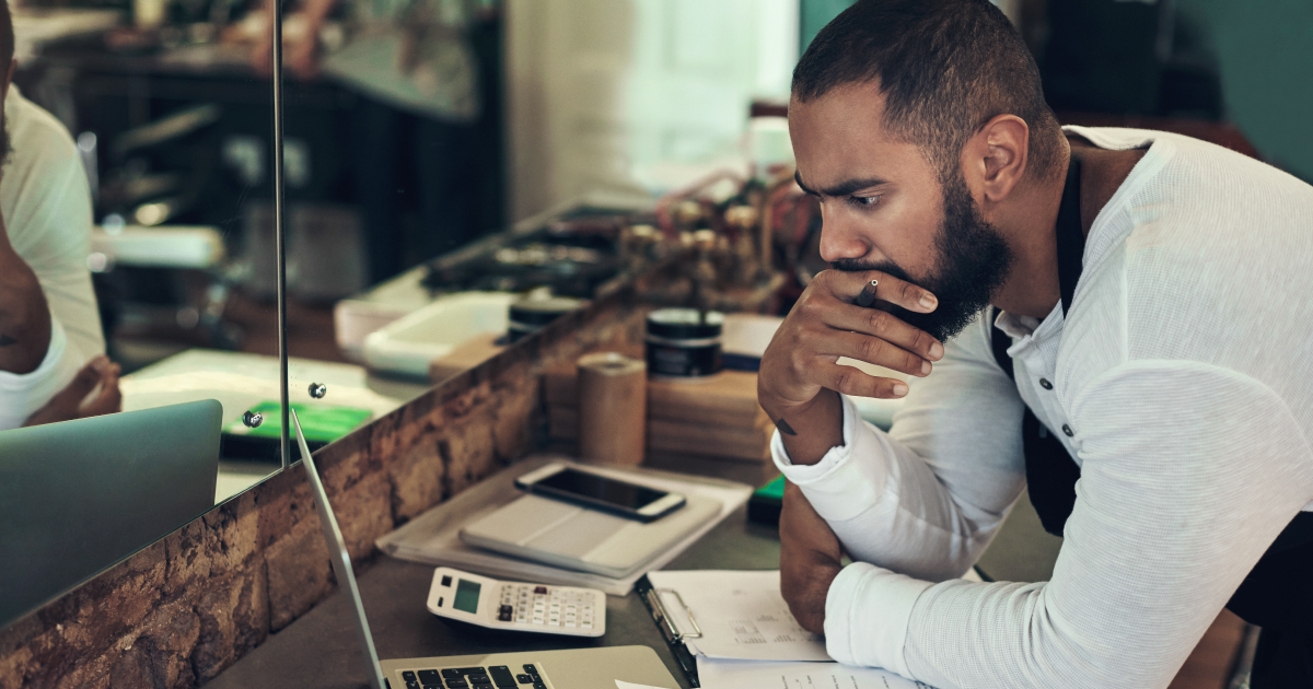 A small business owner sits at his counter, leaning over a laptop with a concerned expression, representing the financial threat that payment fraud poses to merchants