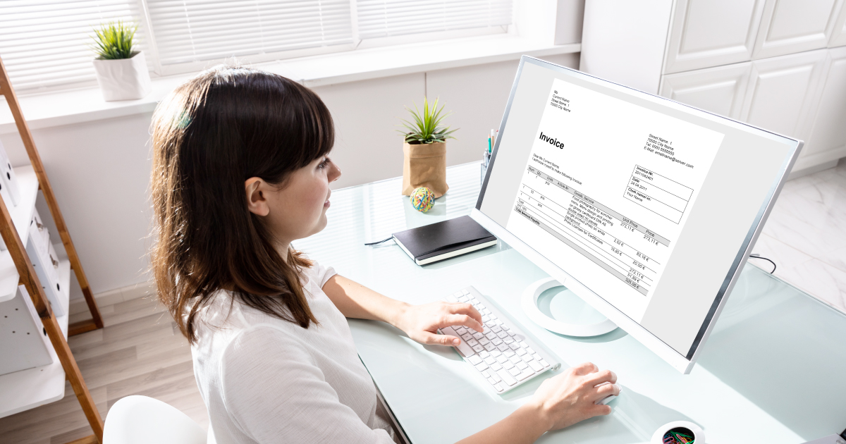 A woman sits at a desktop computer reviewing a digital invoice, representing the invoicing and billing challenges that service-based merchants face every day.