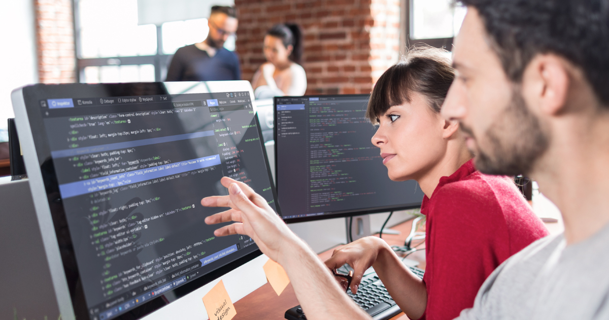 Two software developers collaborate at a desk reviewing code across multiple monitors in a modern open-plan office.