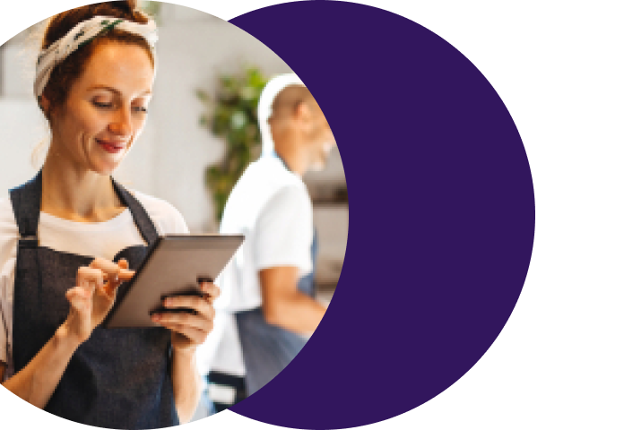Smiling merchant business owner in an apron reviewing onboarding progress on a tablet, representing the streamlined path from merchant sign-up to payment acceptance enabled by Merchant Central's combined web forms, automated workflows and eSignature tools.