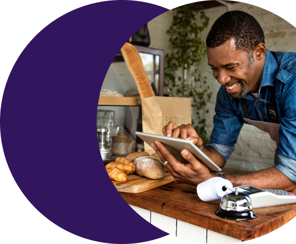Smiling merchant business owner in an apron reviewing a tablet at a food counter, representing the faster and simpler payment activation experience delivered through Merchant Central's web form onboarding tools.