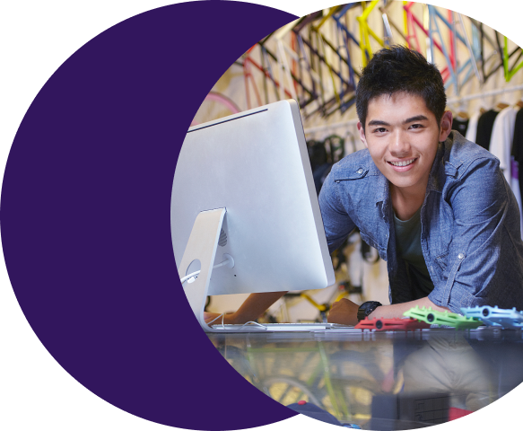 Smiling young man working at a desktop computer in a retail or creative environment, representing a merchant or support agent using Merchant Central's helpdesk collaboration tools to manage and respond to support requests.