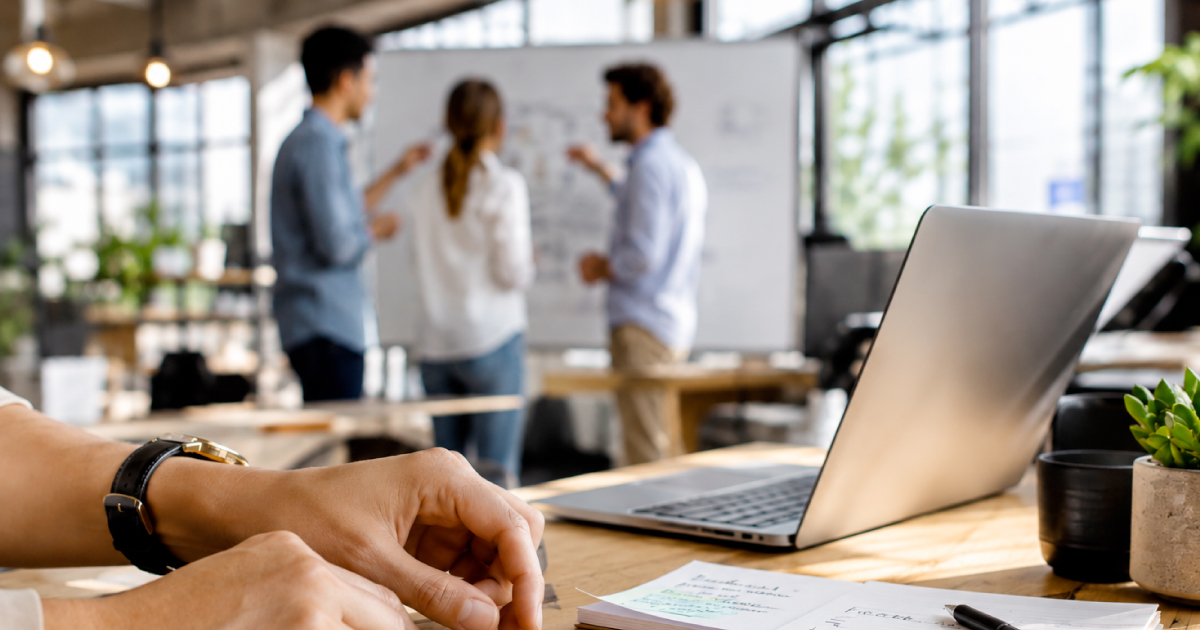 Three colleagues stand at a whiteboard discussing a diagram while a fourth person works at a laptop in the foreground of a bright, modern office.